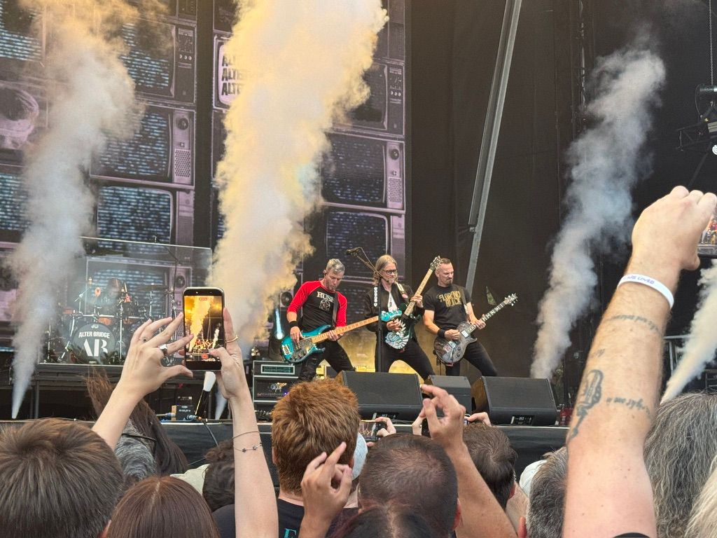 Alter Bridge on stage; the drummer Scott Phillips is in the background while the bassist Brian Marshall, guitarist and vocalist Myles Kennedy, and guitarist Mark Tremonti are on the front of the stage wielding their guitars.