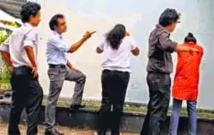 Recreation of a hazing scene: three men are standing behind and making threatening gestures at two women, who seem to be crying.