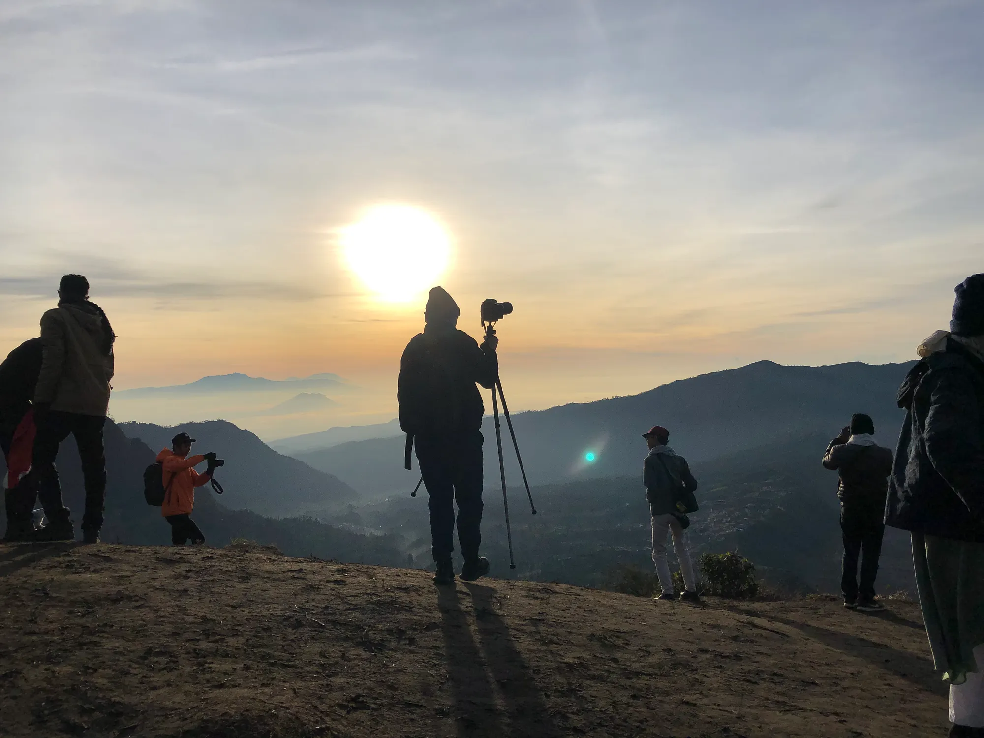 A man standing with a large tripod on a hill, while the sun rises in the background