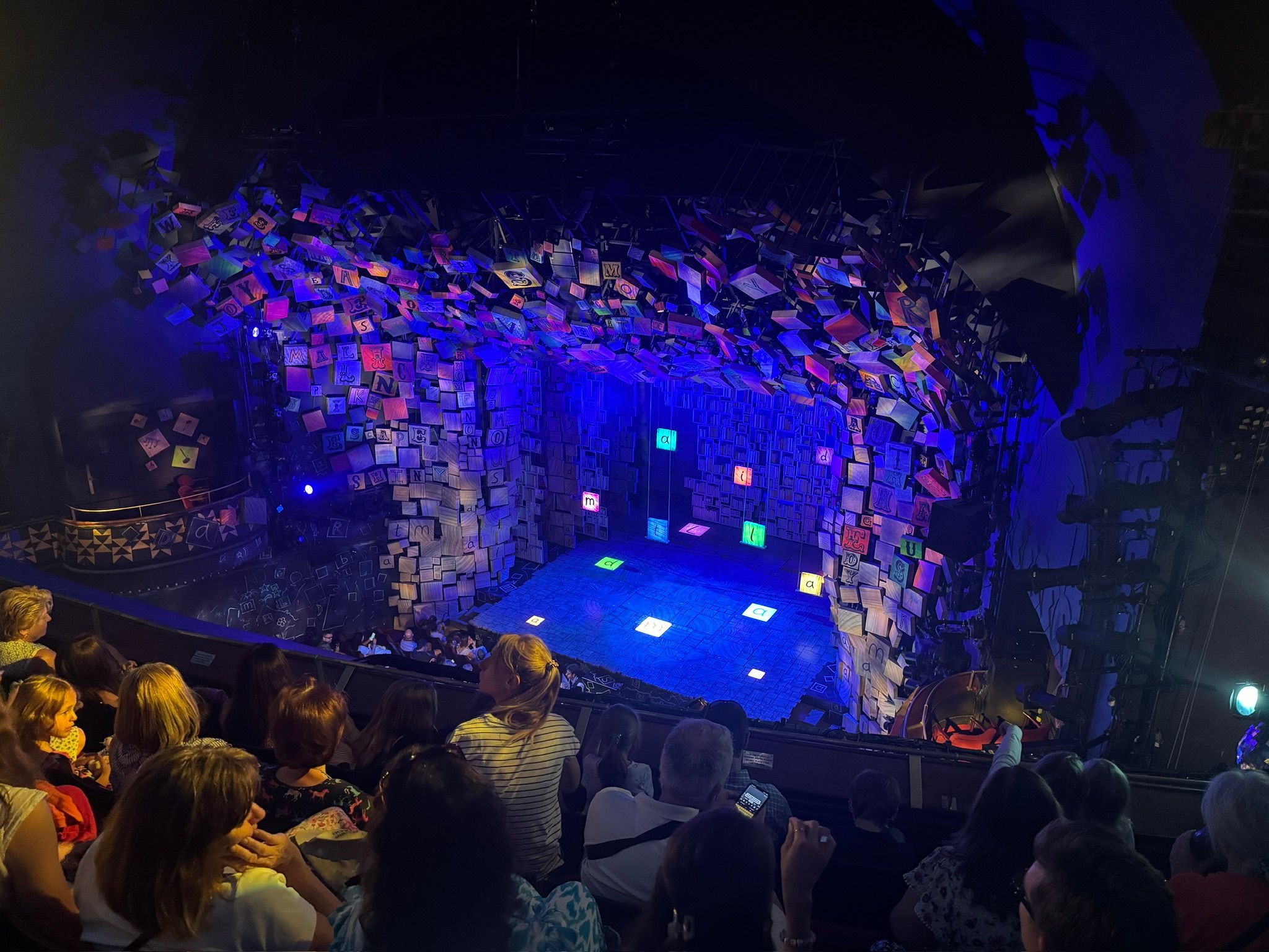 The stage of the Cambridge Theatre, illuminated in multi-cloured lights and decorated with a large set piece resembling scrabble tiles bursting out of all the sides of the stage