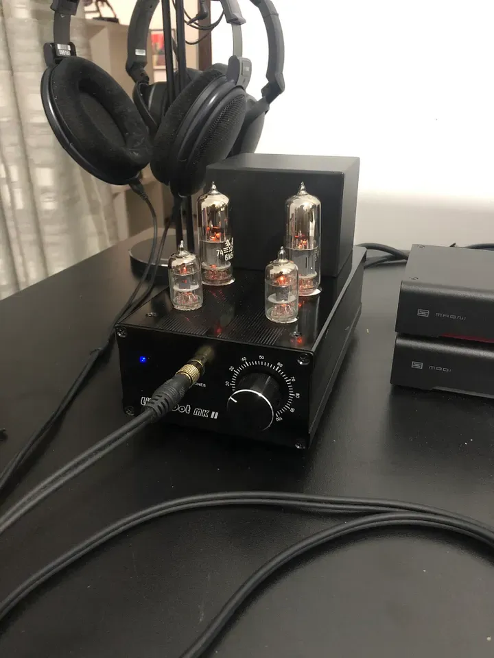 The amplifier, turned on, indicated by a blue light on the front panel. A pair of large headphones are plugged into it.