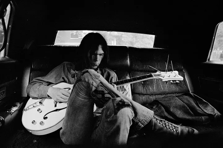 Neil Young in the back seat of a car, with one of his white Gretsch hollow-body guitars