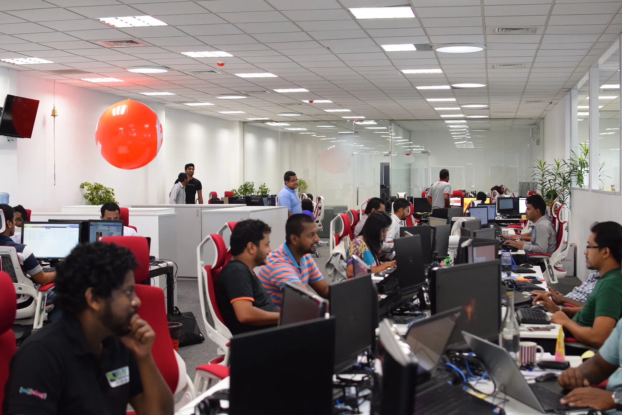 An office, with people seated in two rows of desks, on red chairs.