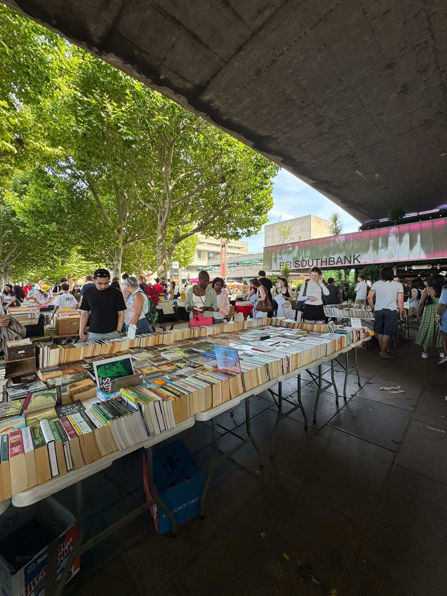 Rows of book set up on tables and people browsing through them. Overhead, the underside of a bridge can be seen.