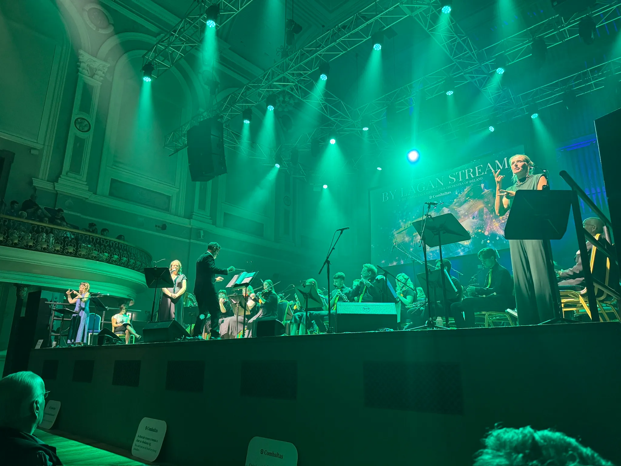 The orchestra performing on stage, seated. The conductor is in the front along with a vocalist, and there are two sign language interpreters on either side of the stage. The stage is lit in green light.