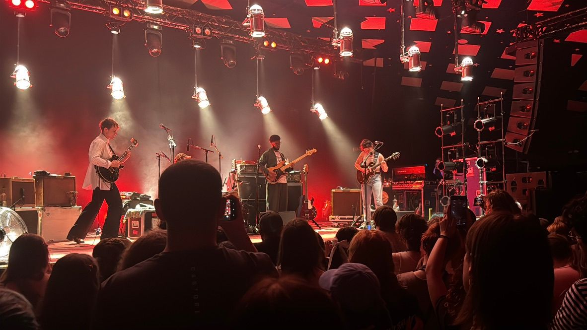 Big Thief on stage illuminated by glowing red lights; guitarist Buck Meek on the left, bassist Joshua Crumbly in the middle and guitaist/vocalist Adrienne Lenker on the right. The drummer is obscured by a head in the audience.