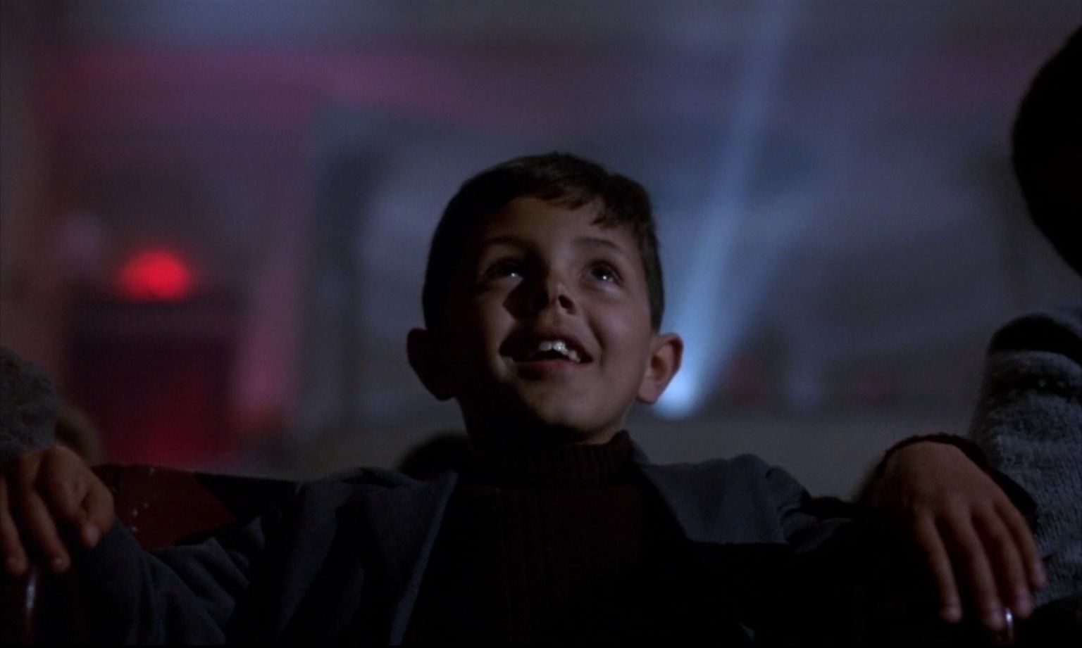 A boy with a smile looking at a cinema screen (the screen is outside the frame but its existence is implied by a beam of projector light behind the boy)