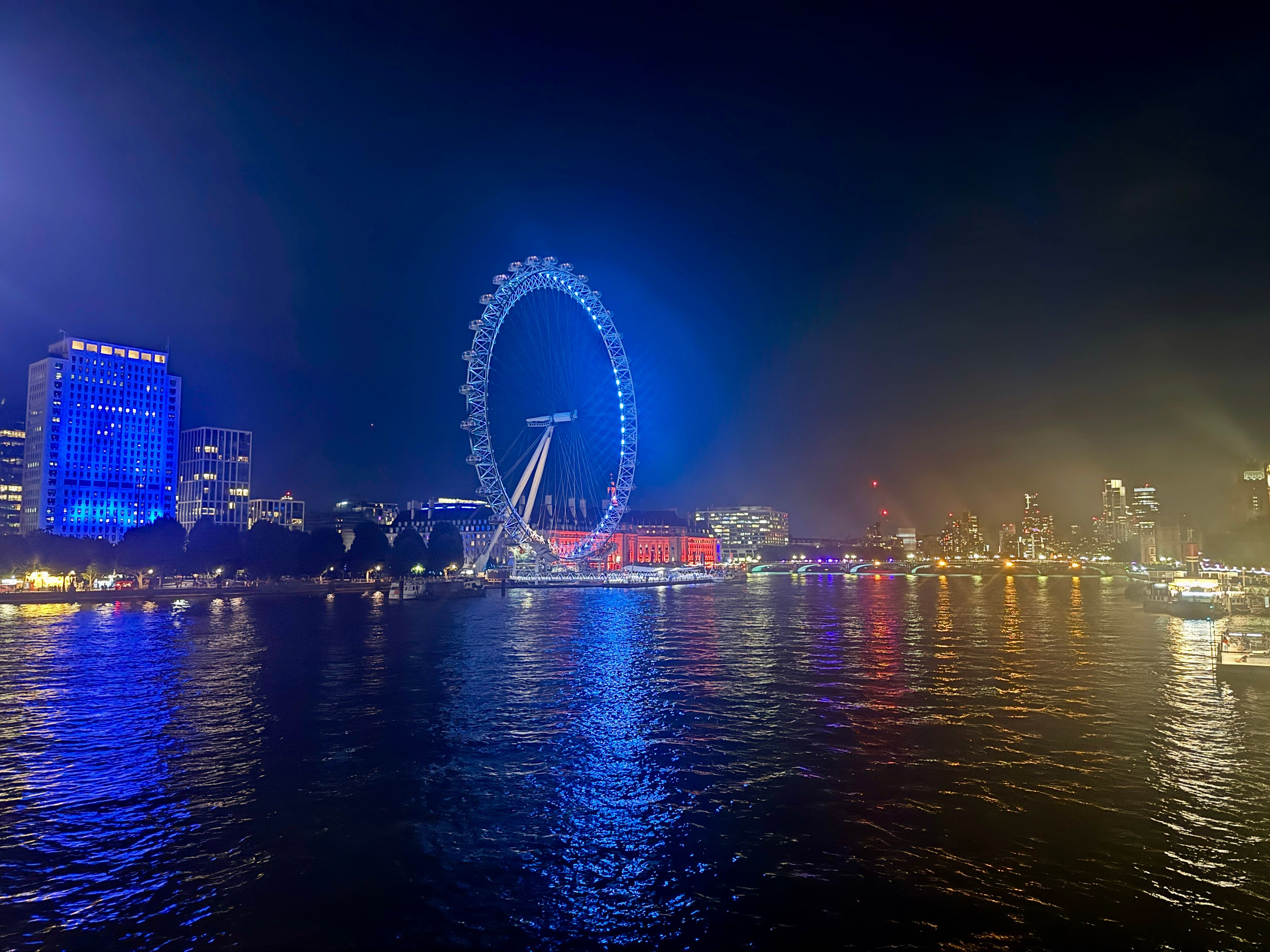 Night views along the Thames river, with the London Eye illuminated in blue, set against the backdrop of bright specs of light emanating from the lamps in the walkways, boats in the river, and buildings on either side.