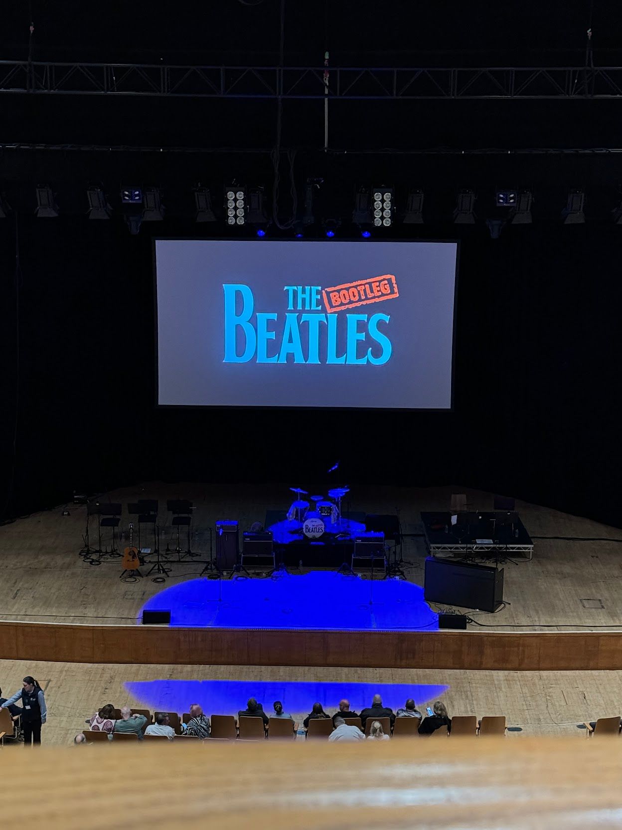 The stage of Waterfrong Hall, with a large screen at the back. The Bootleg Beatles logo is projected on the screen.