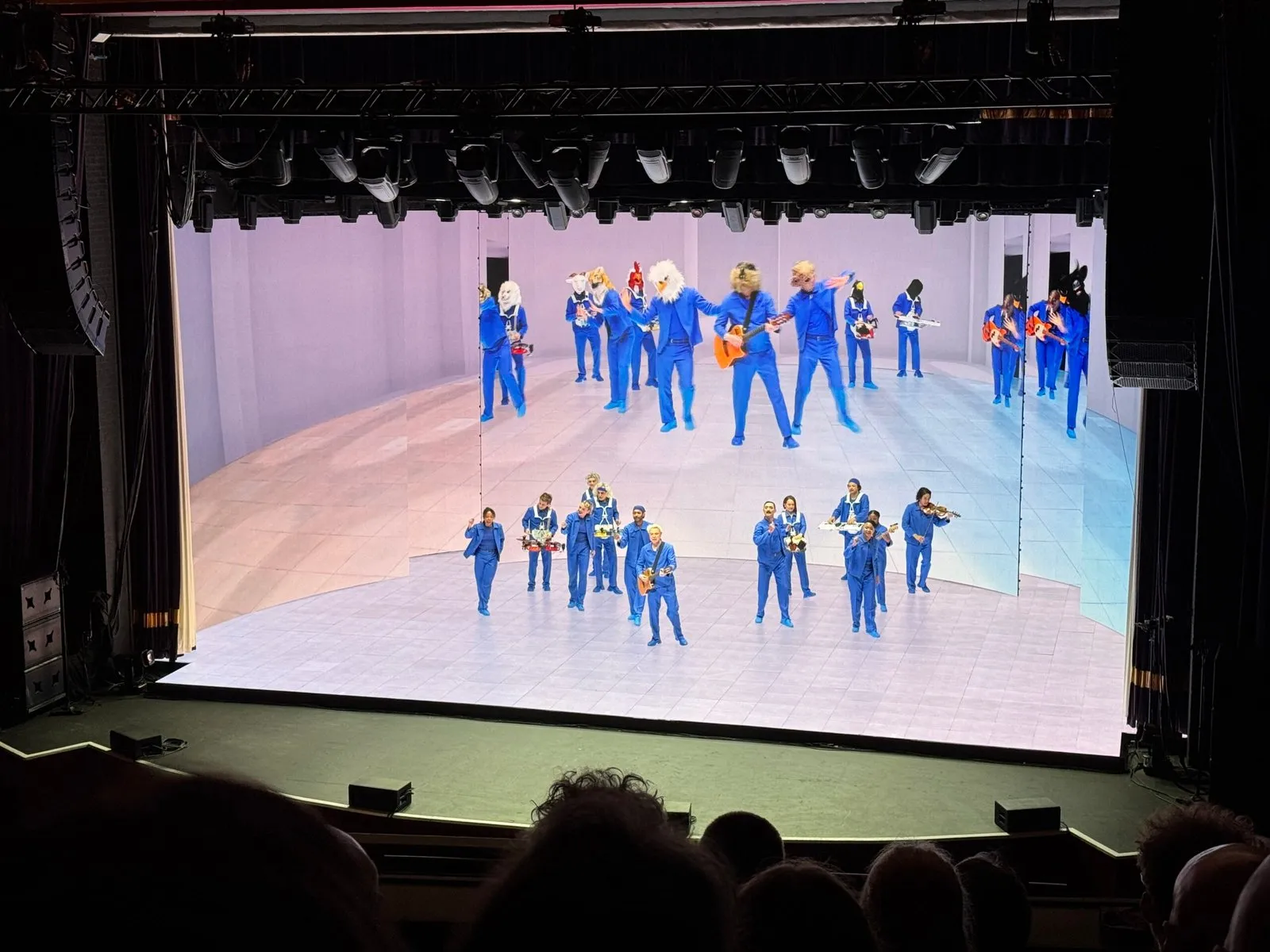 A photo of the performance, with David Byrne and the crew on stage. A large screen behind them is playing a pre-recorded version of the performance that complements the on stage one.