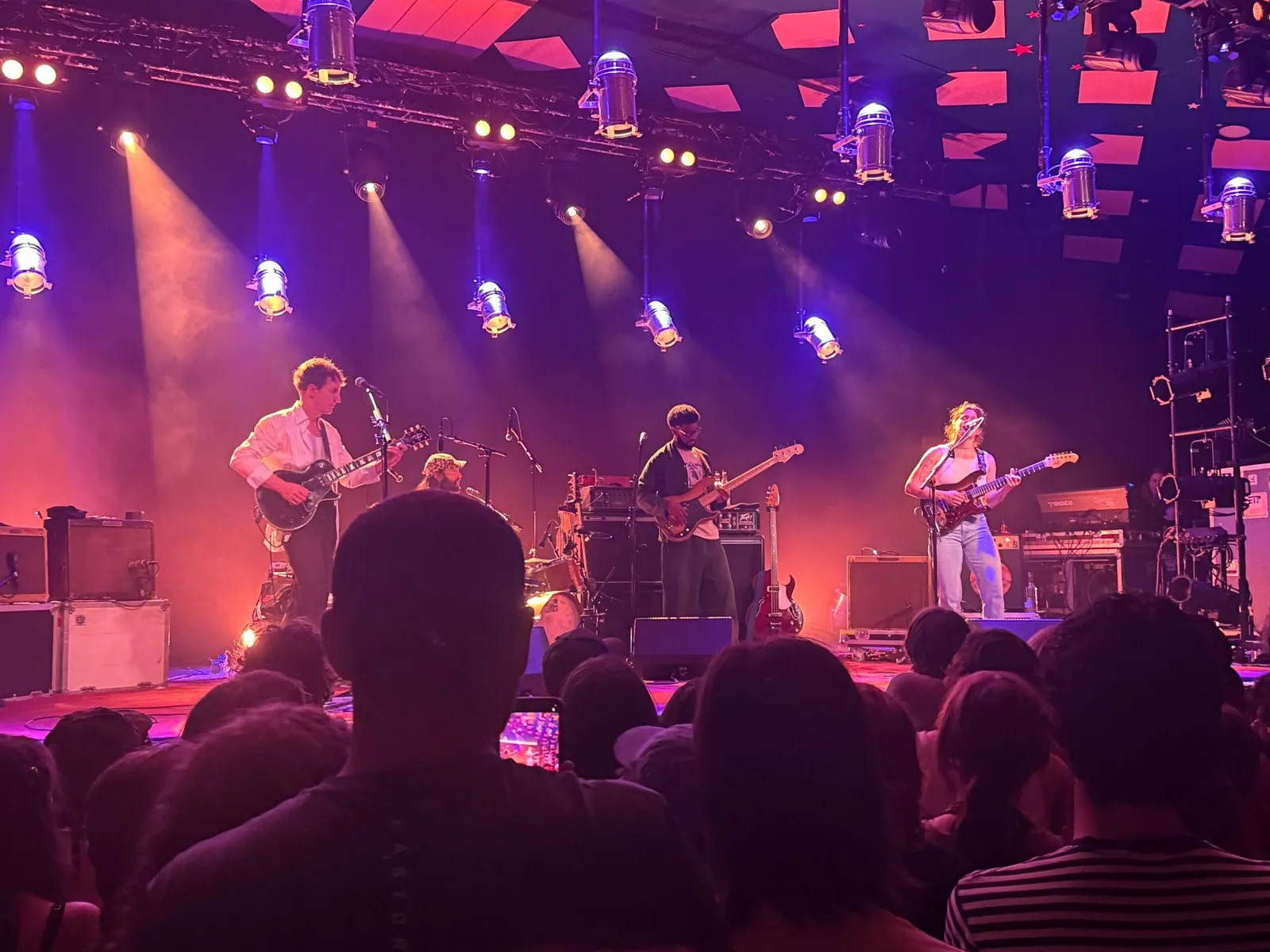 The band on stage, mid-performance. They’re illuminated by soft orange and purple lights while the audience in the foreground looks on.