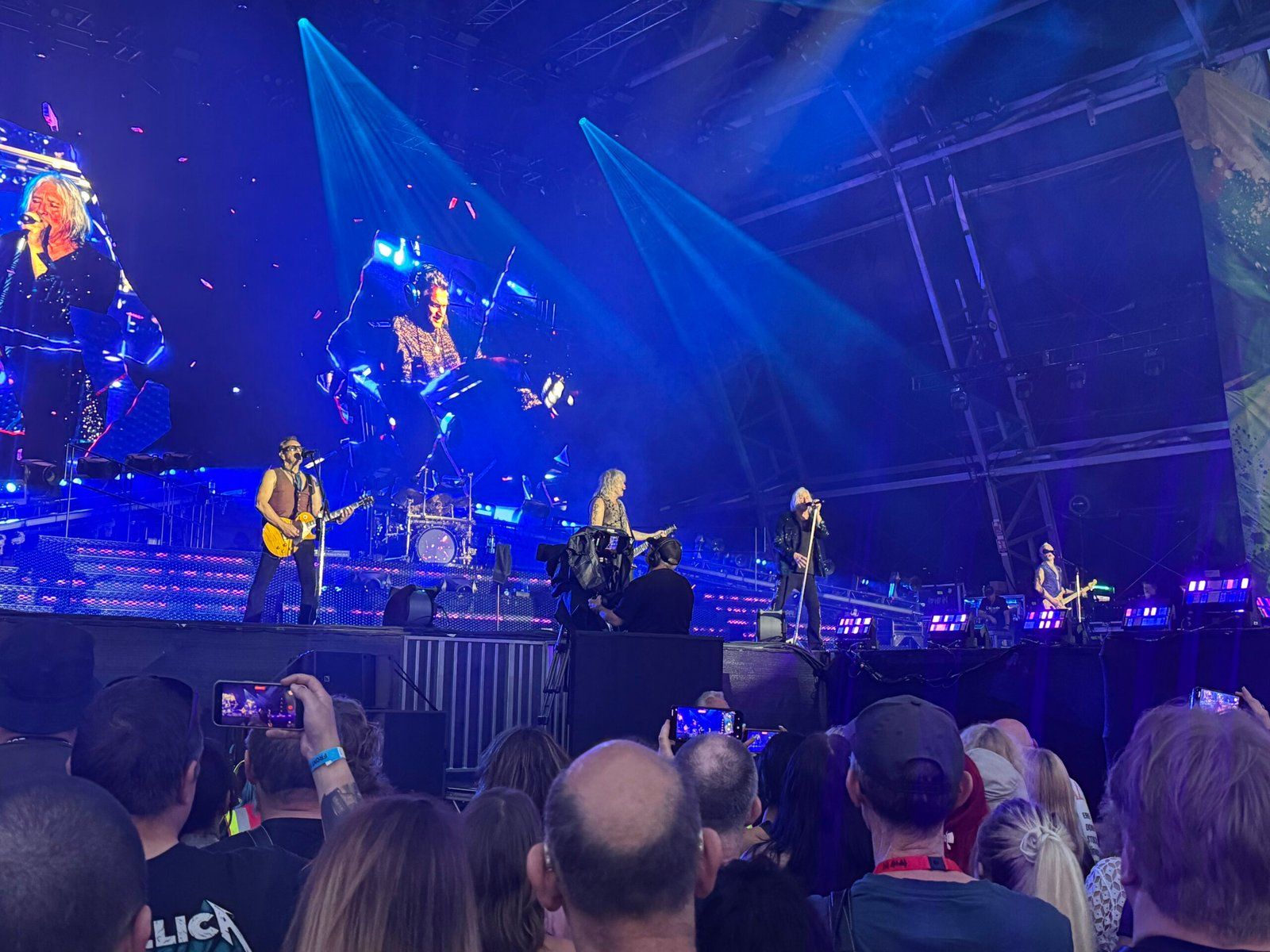 Def Leppard performing on stage. From left to right: Vivian Campbell on guitar, Rick Allen on drums (visible on the large screen at the back), Rick Savage on bass, Joe Elliott on vocals, and Phil Collen on guitar. The stage is washed in blue light.
