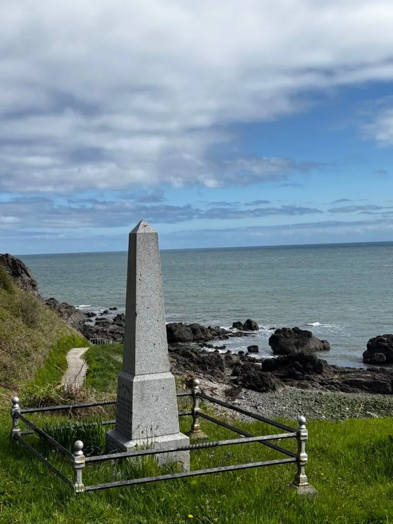 An obelist erected on the coast near the shore. The craggy shoreline can be seen in the background, and a section of the path can be seen to the left of the photo.