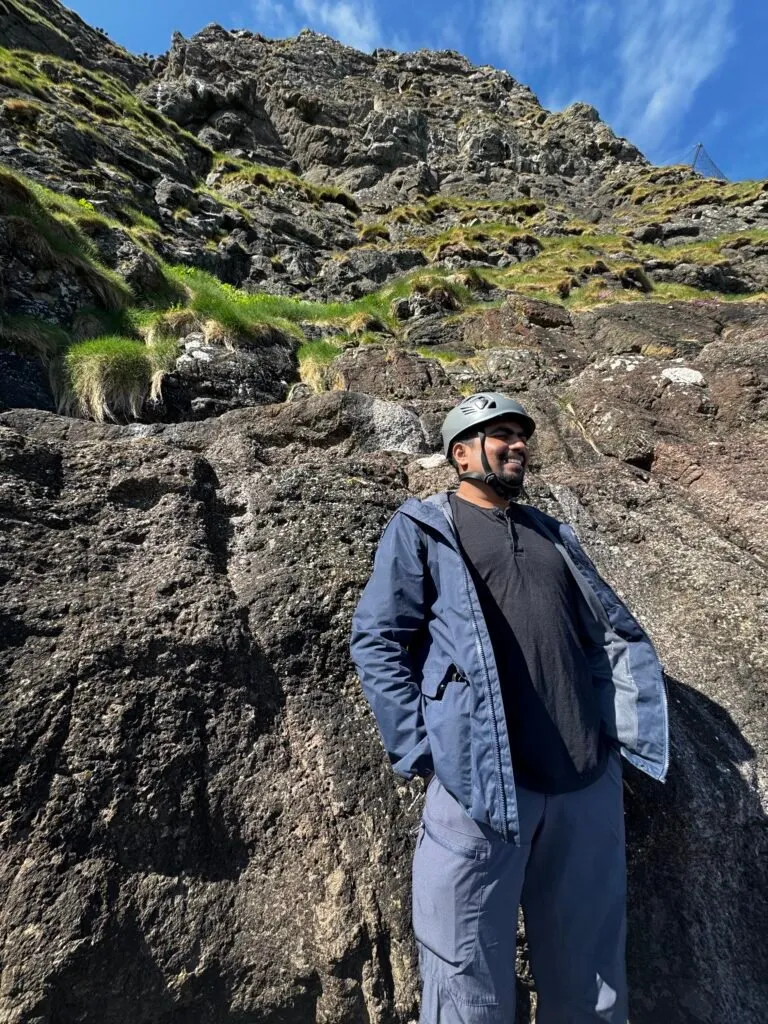 Me wearing a blue coat, blue hiking trousers and a hard hat standing with my back to a cliff.