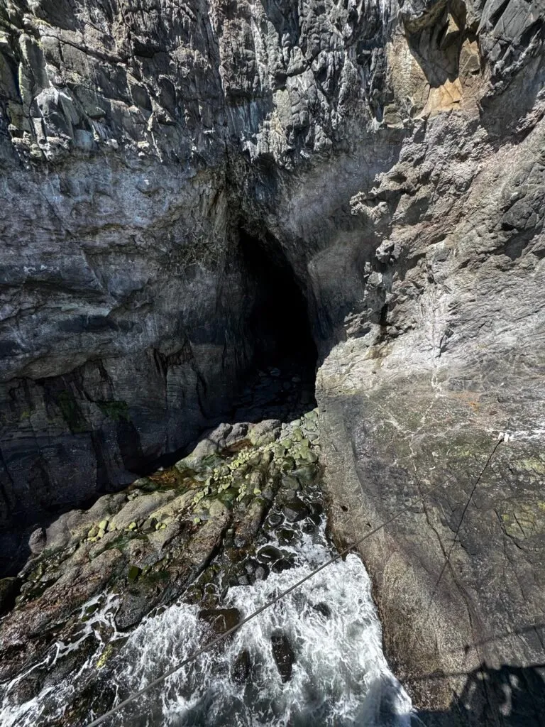 The dark opening of a cave, with a craggy landing in the foreground. Foamy seawater is creeping towards the cave opening.