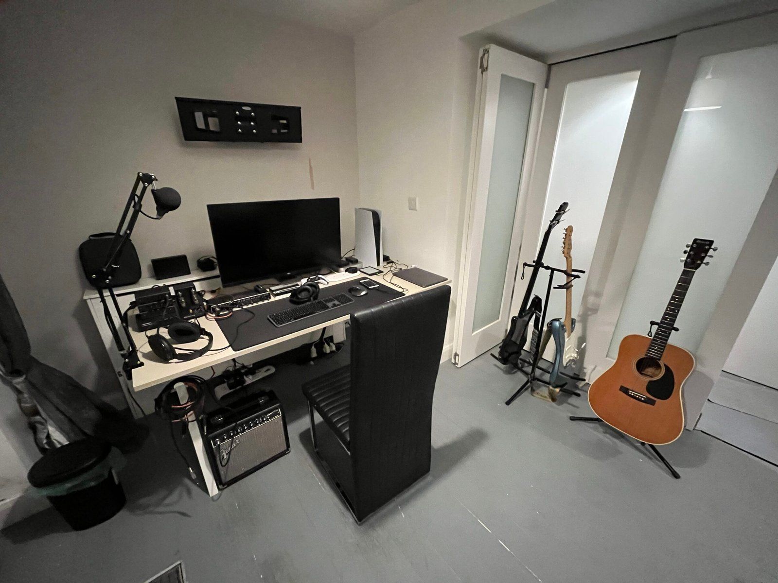 Desk setup featuring monitor, PS5, mic, wireless keyboard and mouse, headphone setup and other peripherals. 3 guitars are resting on stands to the right of the desk.