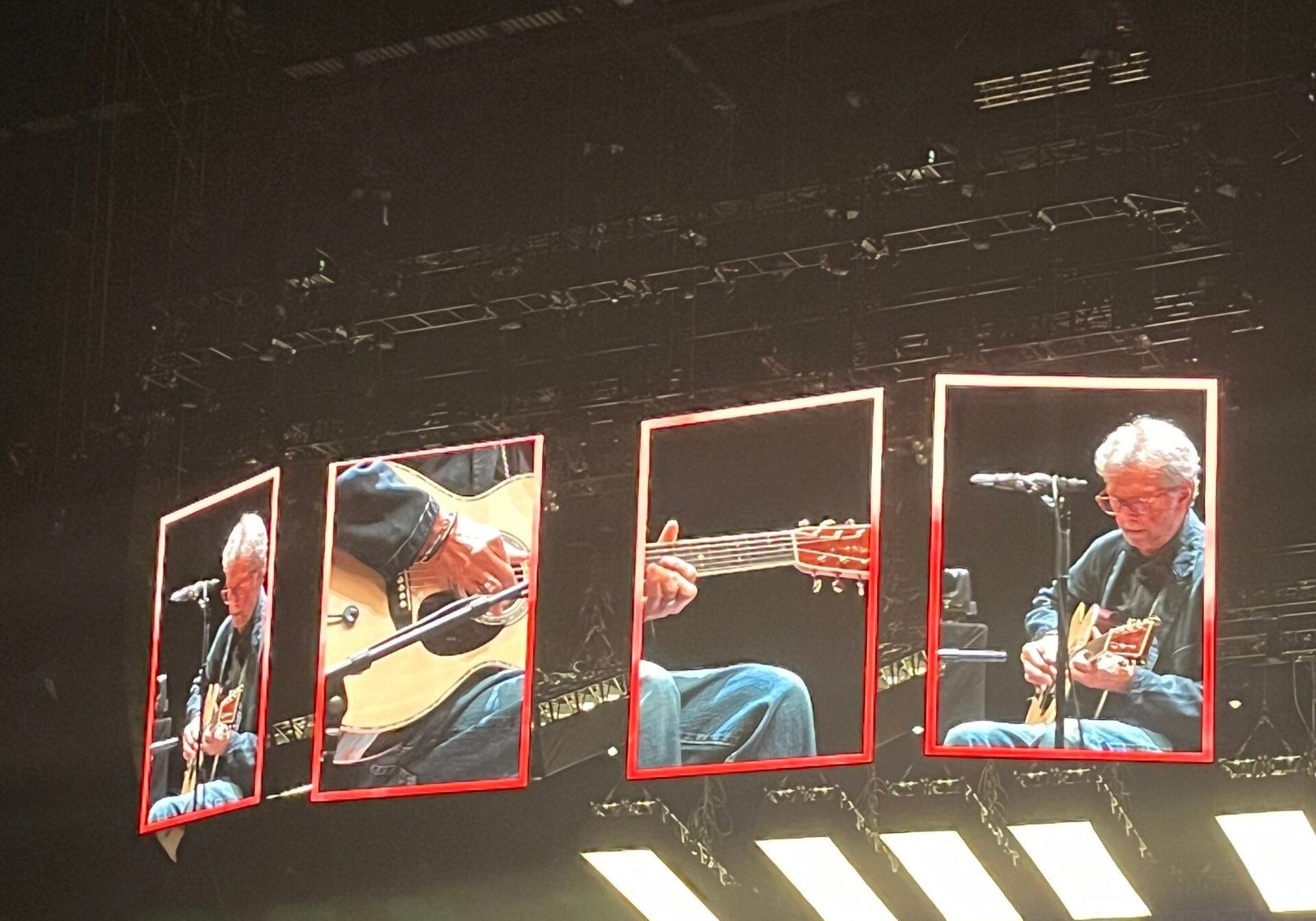 Eric Clapton playing an acoustic guitar on stage as seen through large overhead screens