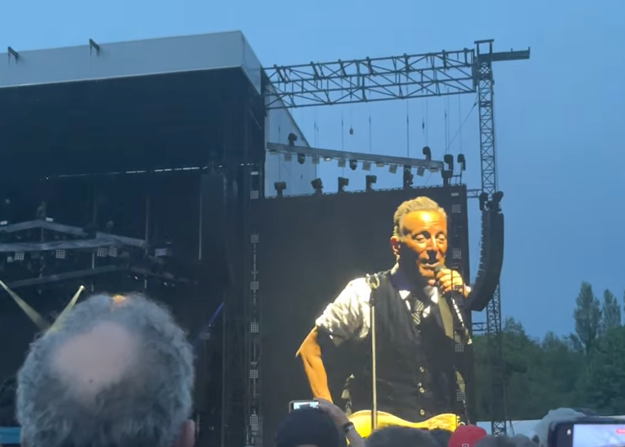 Bruce singing on stage, mic in hand, as seen through a large screen on the right hand side of the stage