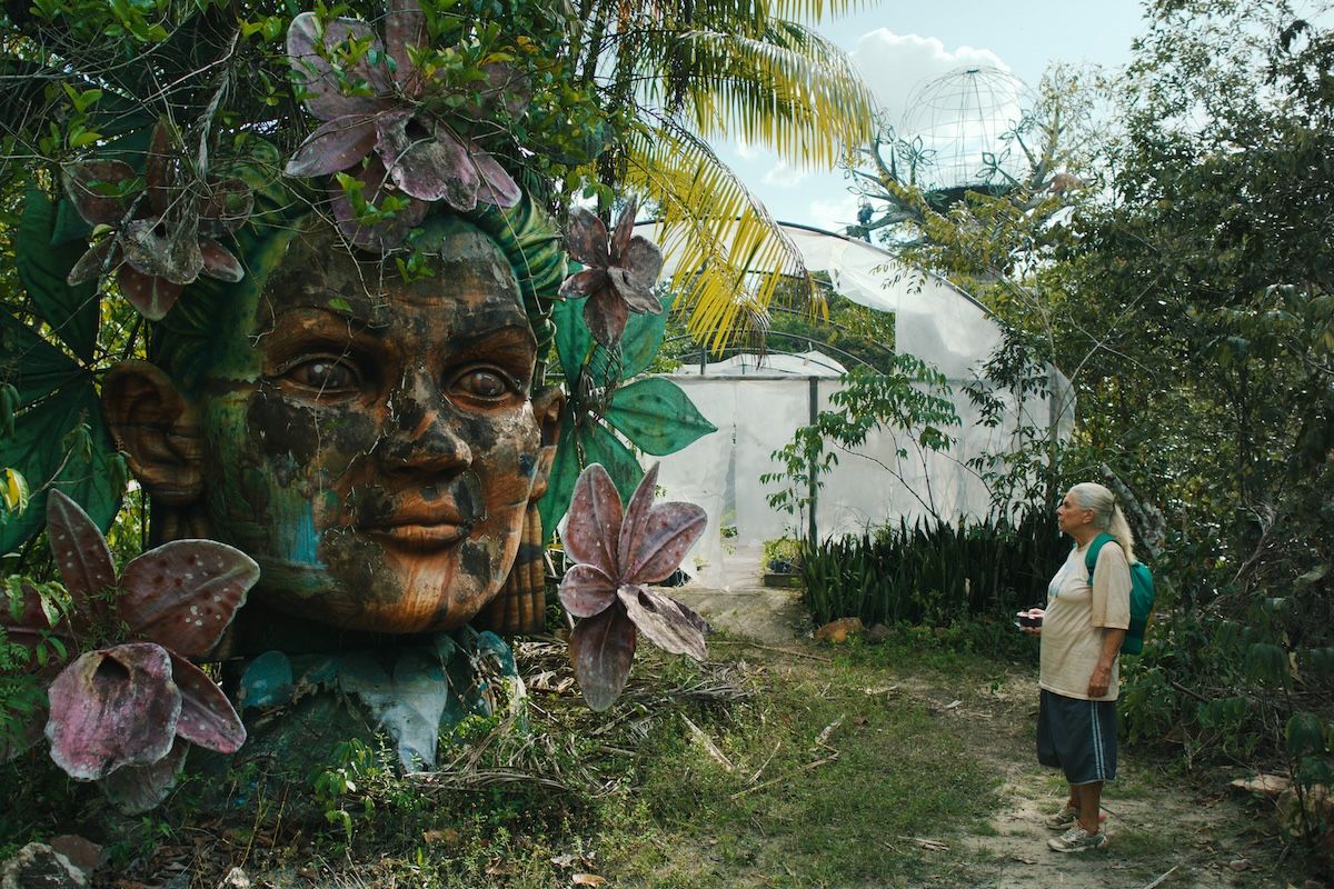 A still from The Blue Trail (2025), where the main character is standing next to a large sculpted head overgrown with foliage