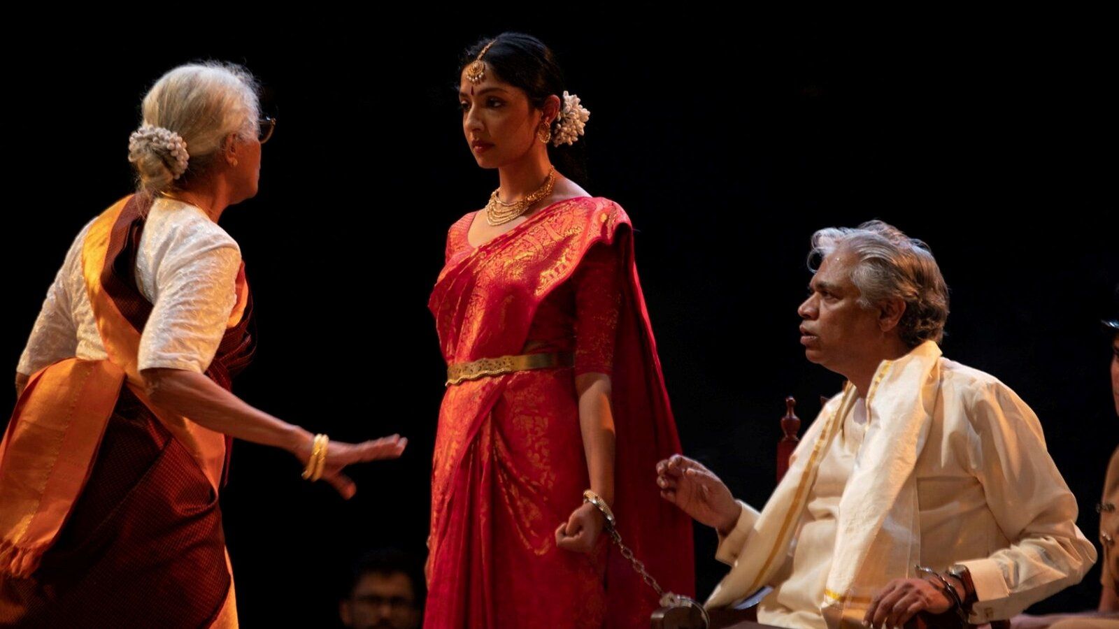 A scene from the play where a woman, dressed in a white and orange saree, is talking to a woman dressed in a bright red saree while a man in a cream top looks on