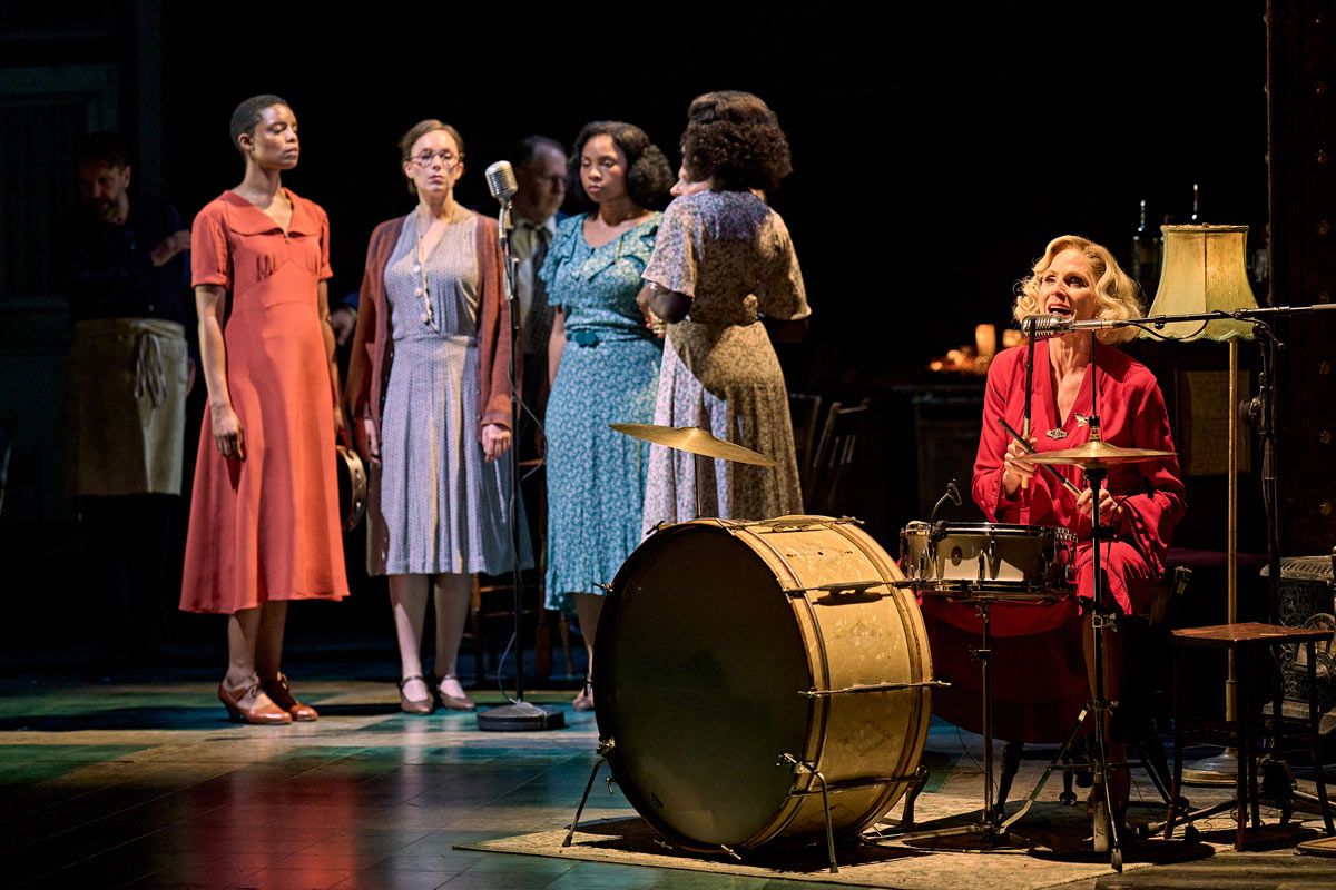 A promotional still from the show; some performers are gathered around a mic on the left of the photo, while one is sitting at a drumset on the right