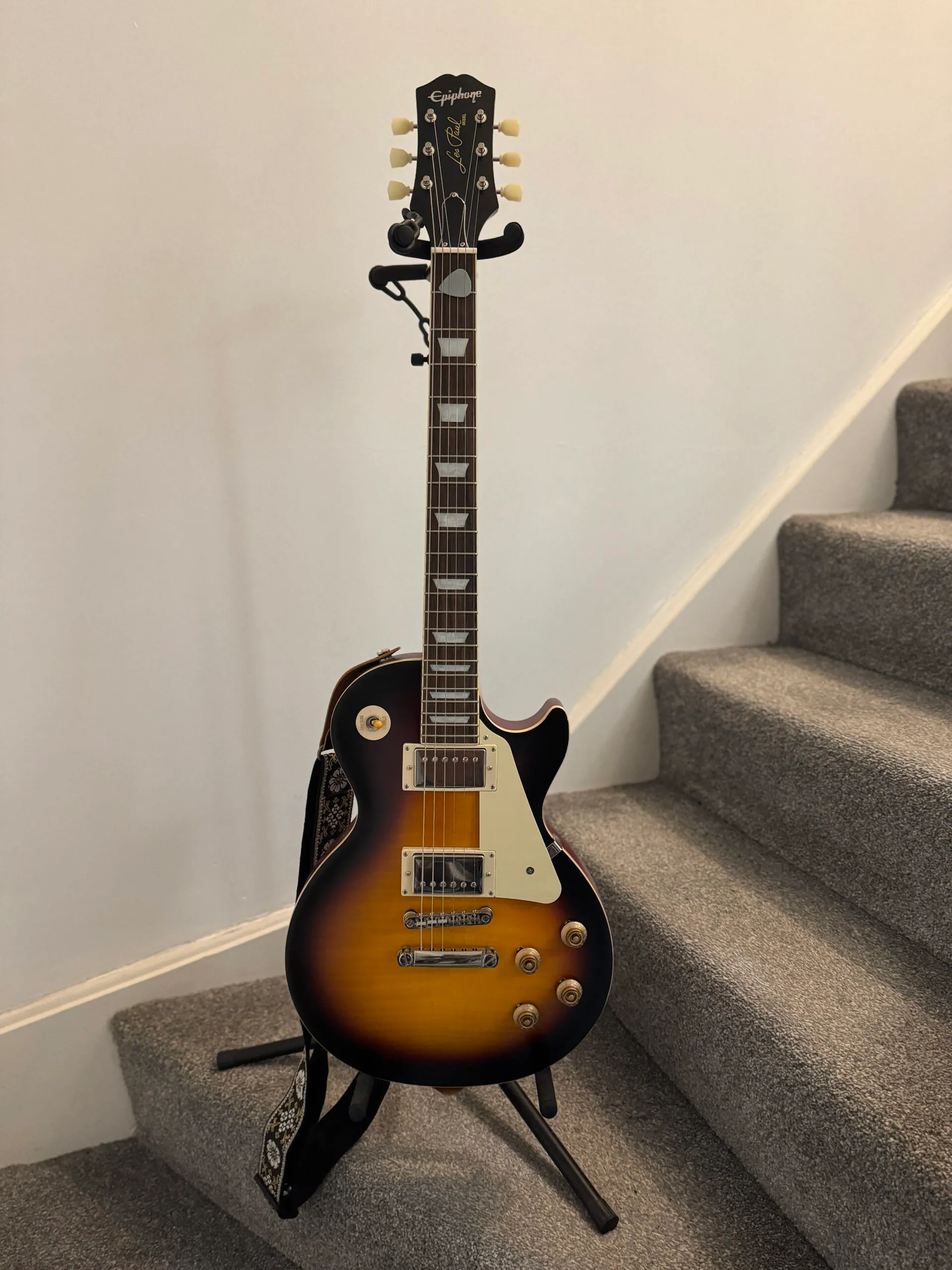 A Les Paul guitar resting on a stand. It has a dark-coloured Laurel neck and a black-to-orange gradient body.