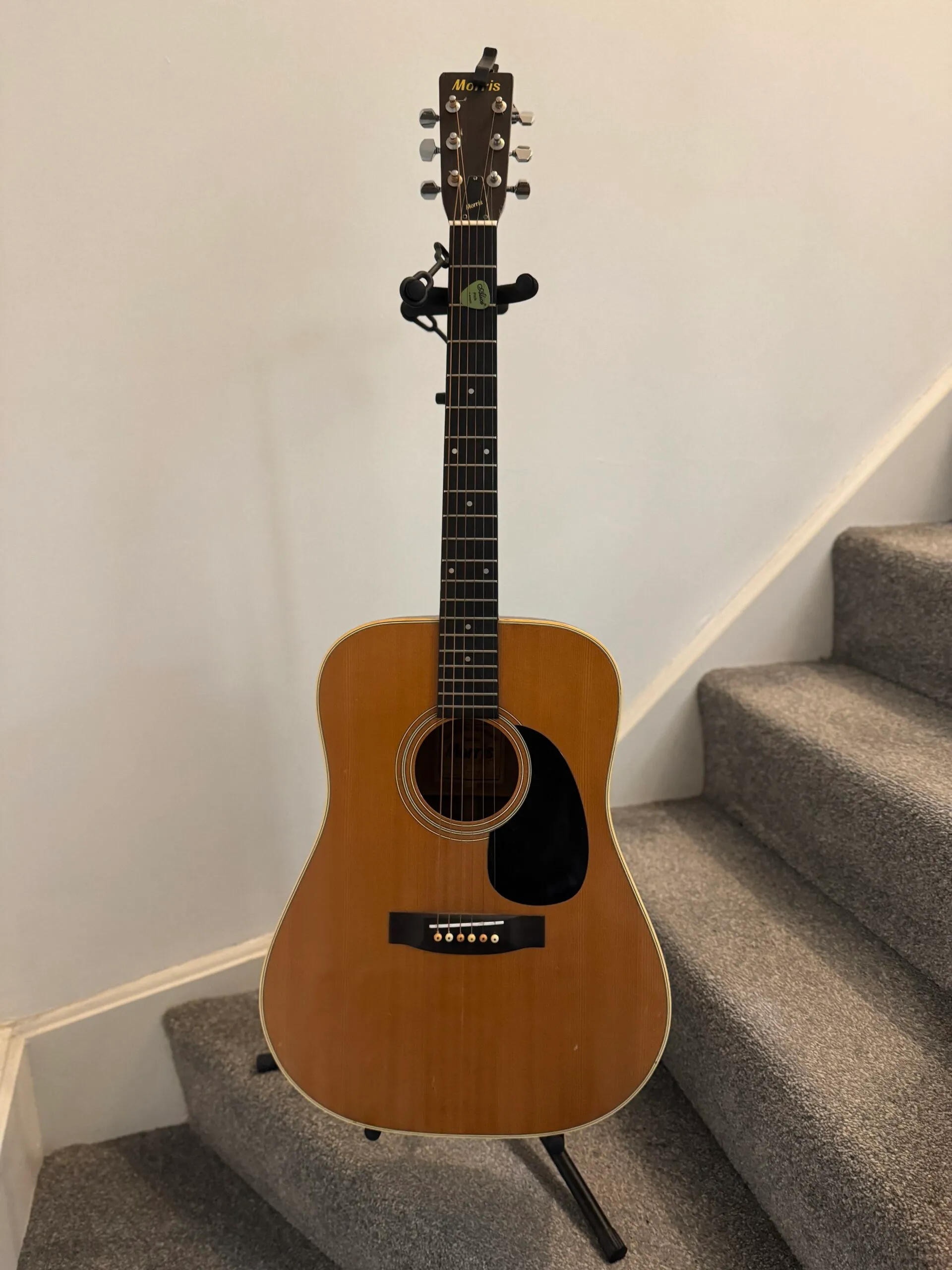 An acoustic guitar resting on a stand. It has a dark-coloured Rosewood neck, a black pickguard, and a natural-wood-coloured Spruce body.