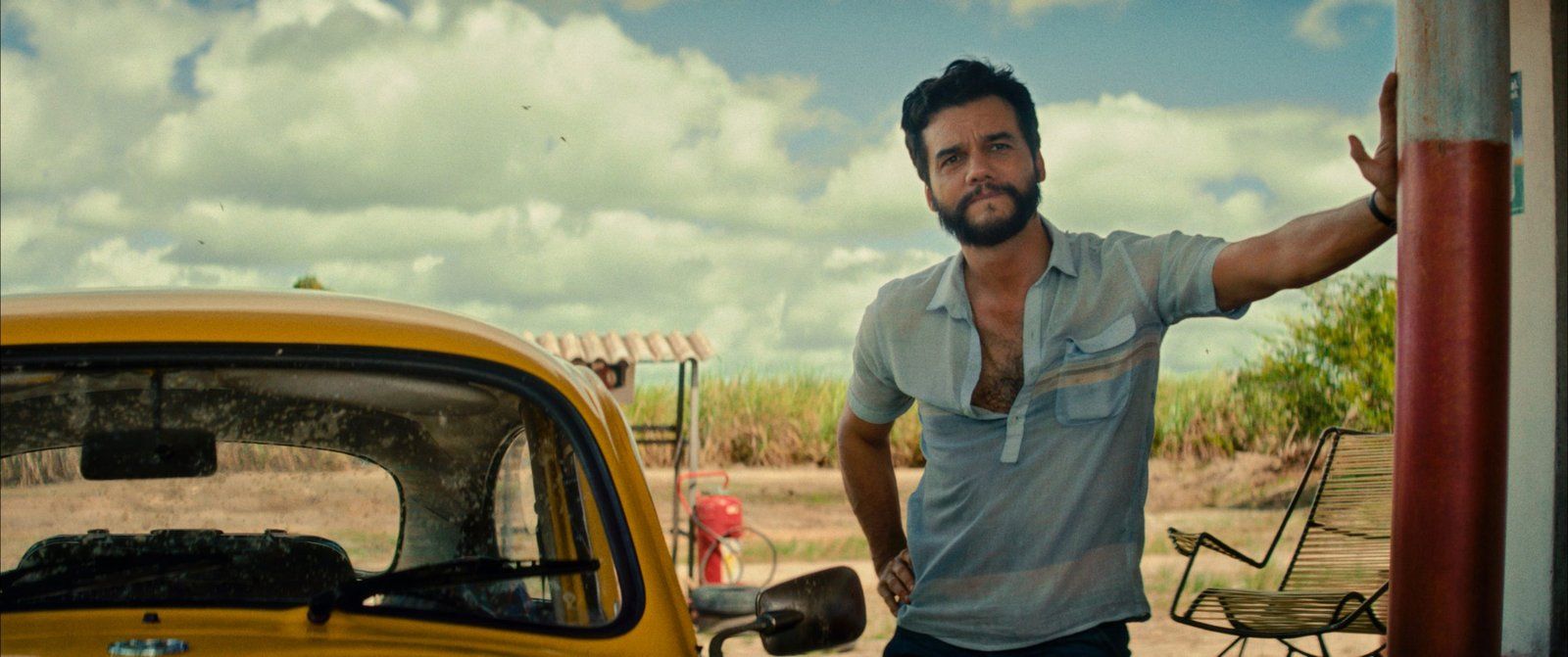 Wagner Moura, who plays Marcelo in The Secret Agent stands next to a yellow car at a gas station in this press still for the film