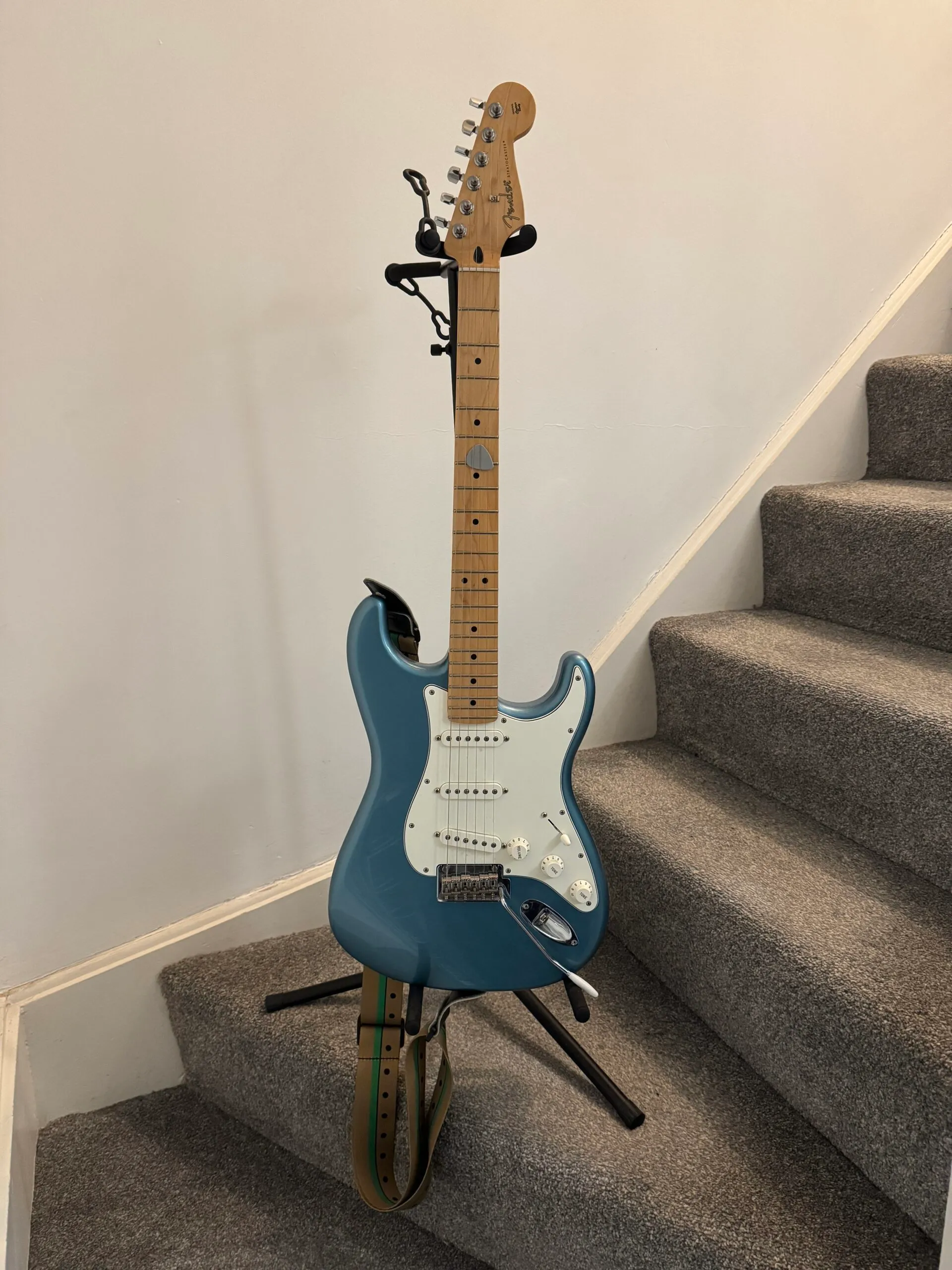 A Fender Stratocaster guitar resting on a stand. It has a light-coloured Maple neck, a turquoise-coloured body, and a white pickguard.