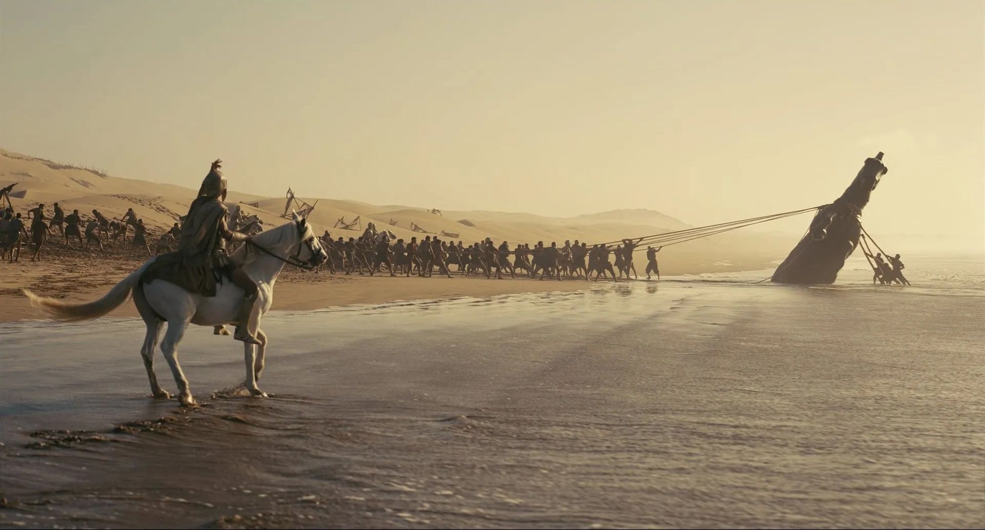 A group of people pulling out a large wooden horse half buried in the sand onto a beach while a horse-mounted soldier looks on