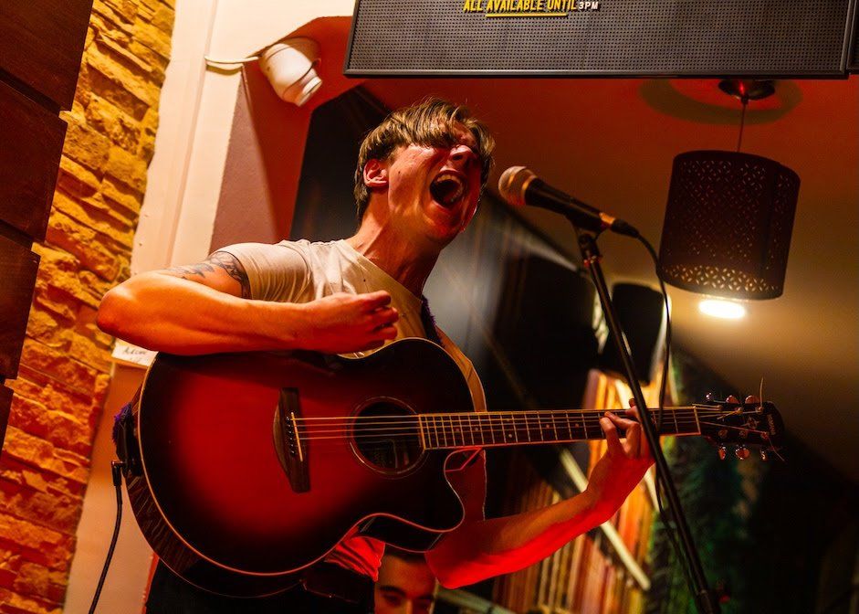 Joseph Toman performing at the event. He is playing an acoustic guitar and screaming into a mic, his mouth open.