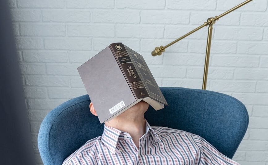 a man in white and blue striped long sleeve shirt sitting on blue armchair with a book on his face