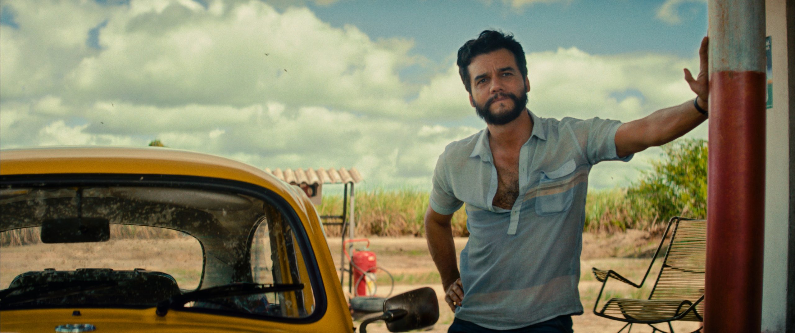 Wagner Moura, who plays Marcelo in The Secret Agent stands next to a yellow car at a gas station in this press still for the film